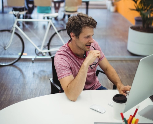 A man in a casual red t-shirt works at a computer in a modern office space with a bicycle in the background.