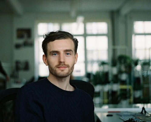 A man with a beard and blue sweater looks directly at the camera in an office setting.