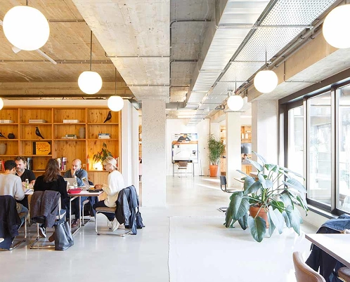 A modern, open-plan office space with people working and a wall of shelves in the background.