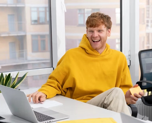 A cheerful man in a yellow hoodie sitting at a desk with a laptop and holding a sticky note.