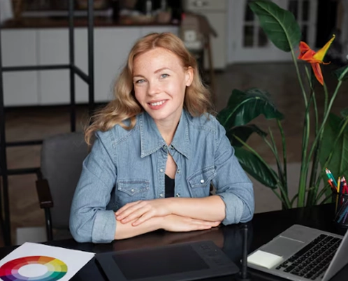 A woman sits at a desk, smiling at the camera, surrounded by a color palette, laptop, and a plant.