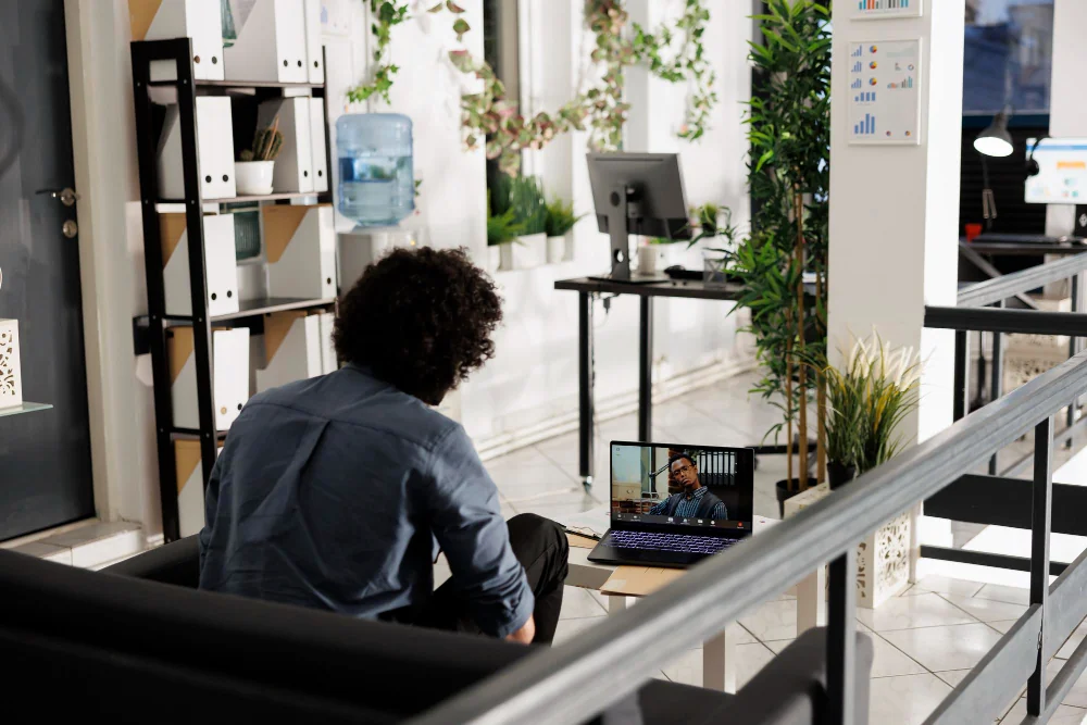 A man sitting on a couch in an office environment, engaged in a video call on a laptop with a coworker.