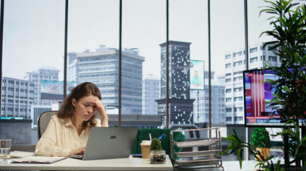 A woman looks stressed while working on her laptop in a modern office with a cityscape view.