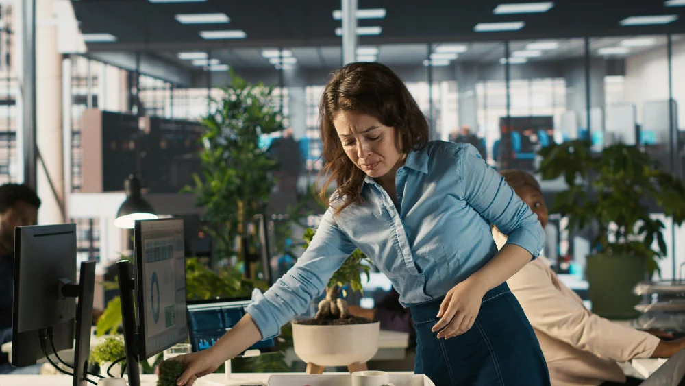A woman looking stressed in an open-plan office environment with greenery around.