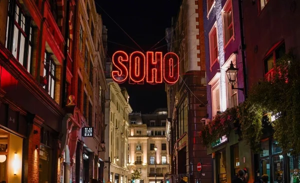 A vibrant street in Soho, London illuminated by a glowing sign at night.
