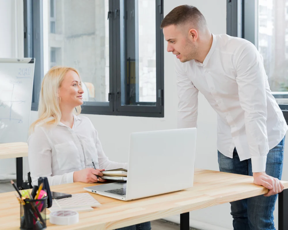 A man and a woman discussing at a desk in a modern office setting.