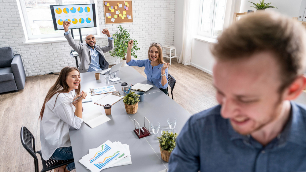 Employees celebrate together in a modern office meeting, with charts and graphs visible in the background.