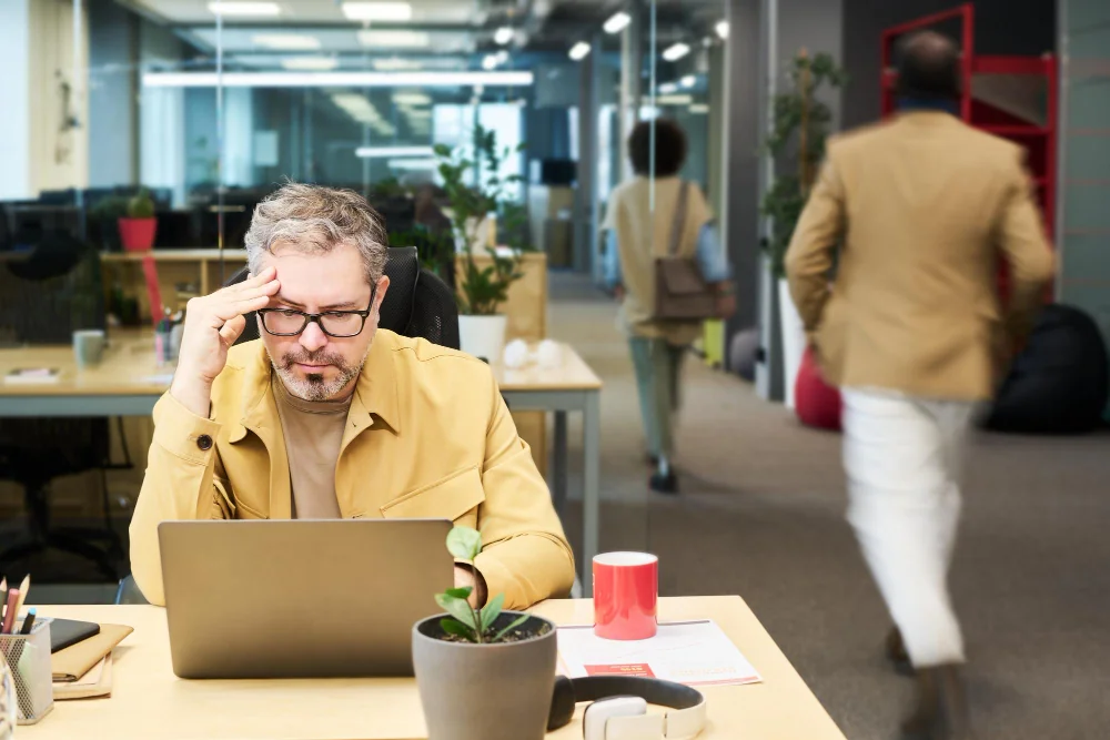 A man in a yellow jacket works intently on a laptop at a partially occupied office.