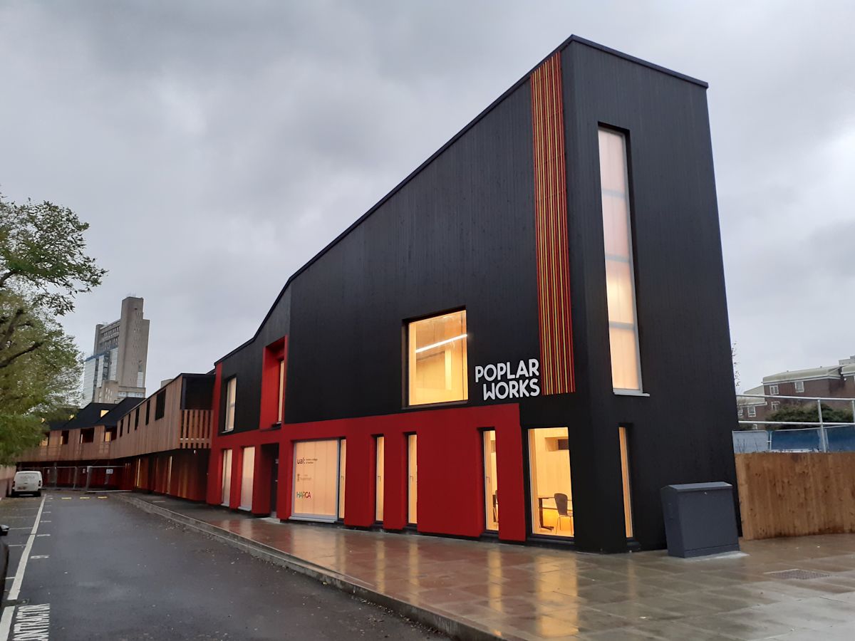 A modern, angular building labeled "Poplar Works" on Abbott Road, London, with a cloudy sky above.