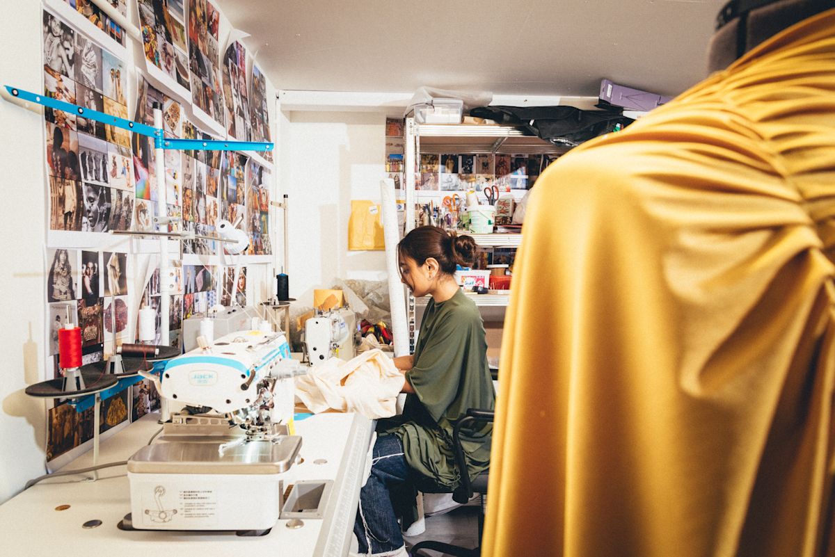 A seamstress works in a fashion studio surrounded by sewing machines and fabric designs.