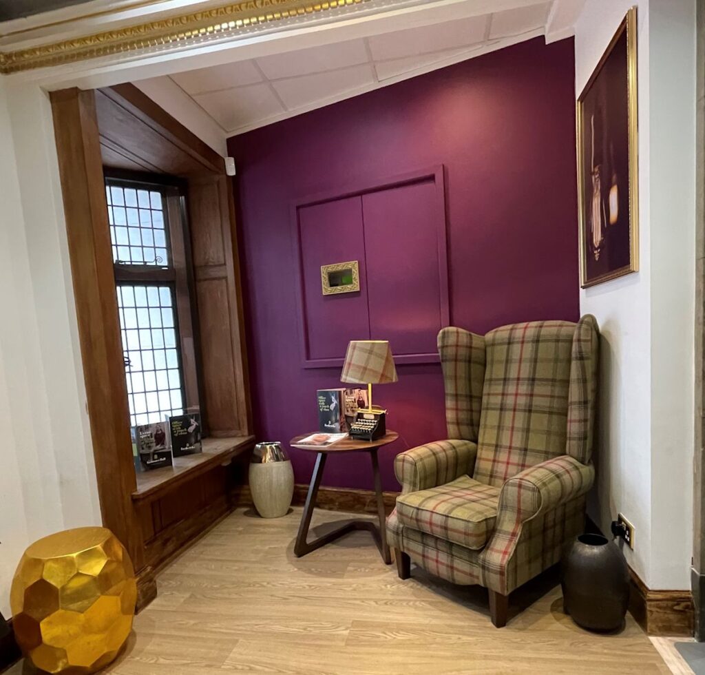 A cozy reading nook with a plaid armchair, small table, and lamp set against a deep purple wall.