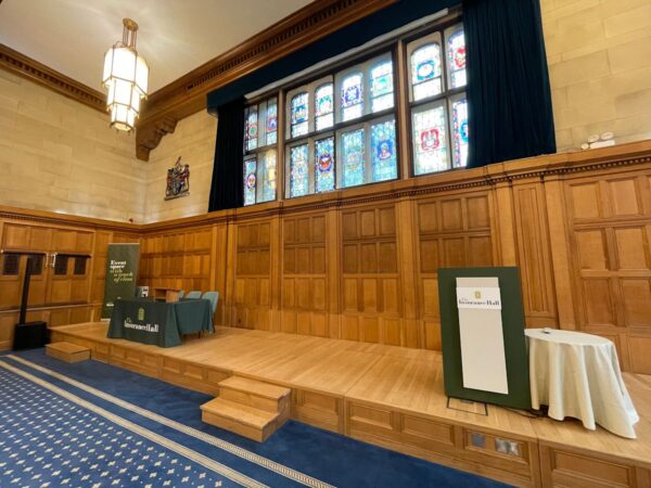 Interior of a wood-paneled hall at Aldermanbury in London, featuring a stage with a lectern and stained glass windows.