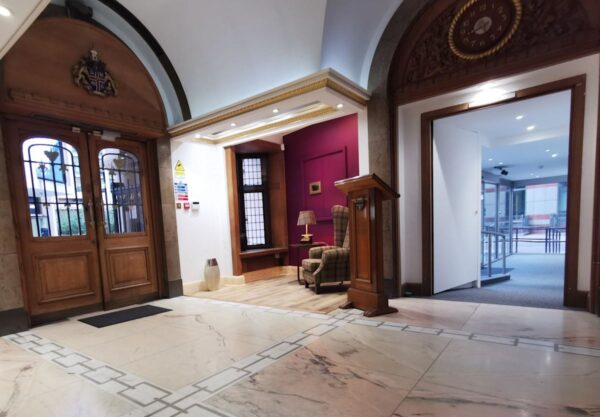 The entrance hall of a London building with marble flooring, wooden doors, and a small seating area.