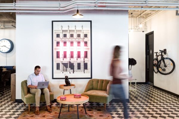 A modern office lounge area with a man working on a laptop, contemporary furniture, wall art, and a bicycle mounted on the wall.