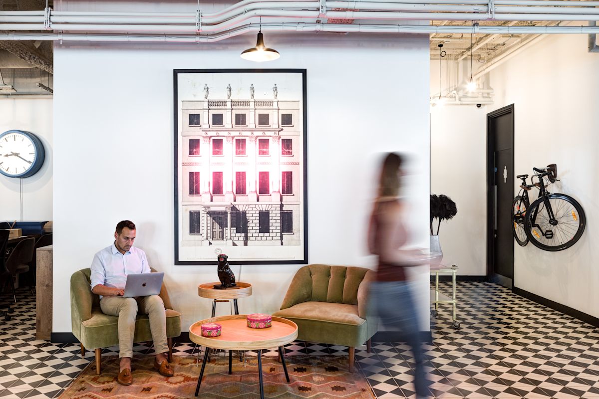 A modern office lounge area with a man working on a laptop, contemporary furniture, wall art, and a bicycle mounted on the wall.