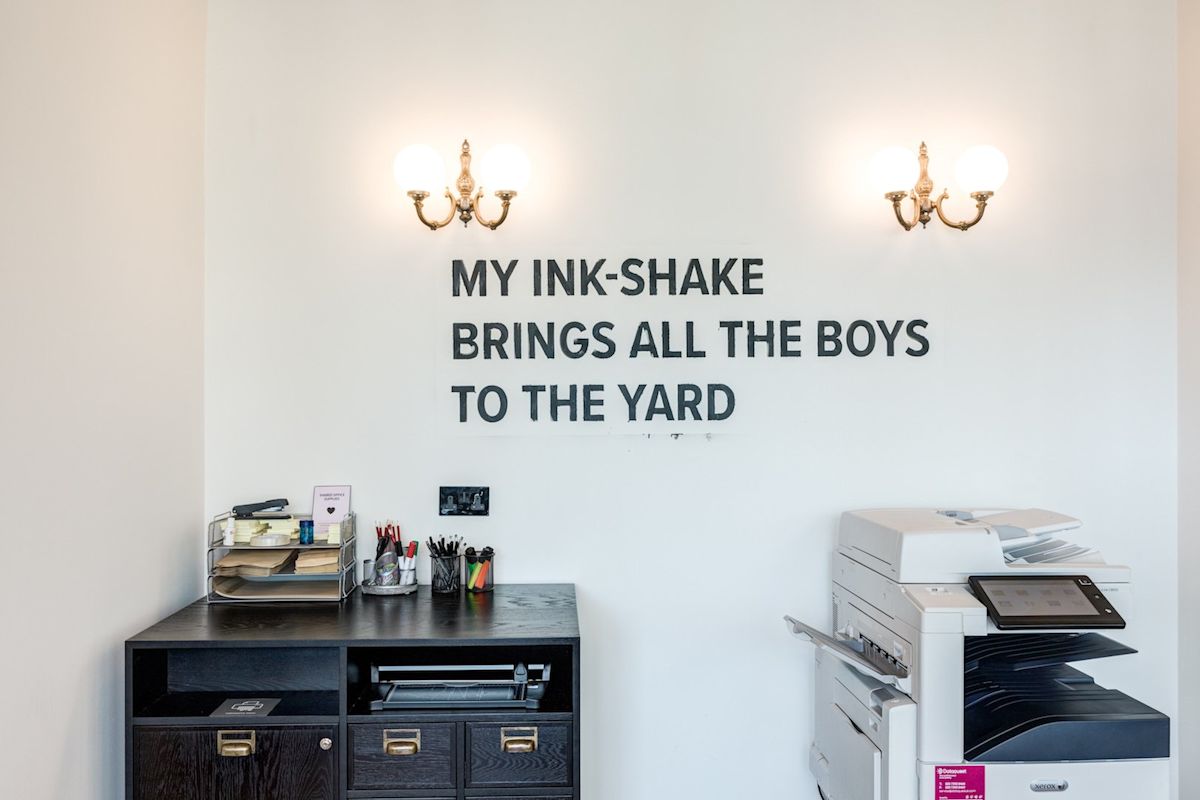 Office corner with a copier and humorous wall text, "My ink-shake brings all the boys to the yard."