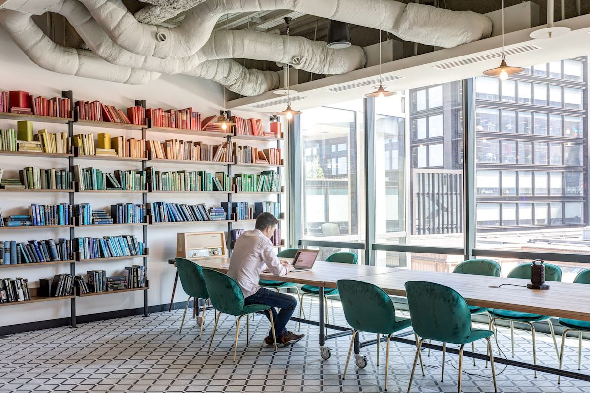 A person works on a laptop in a modern office with colorful bookshelves and large windows.