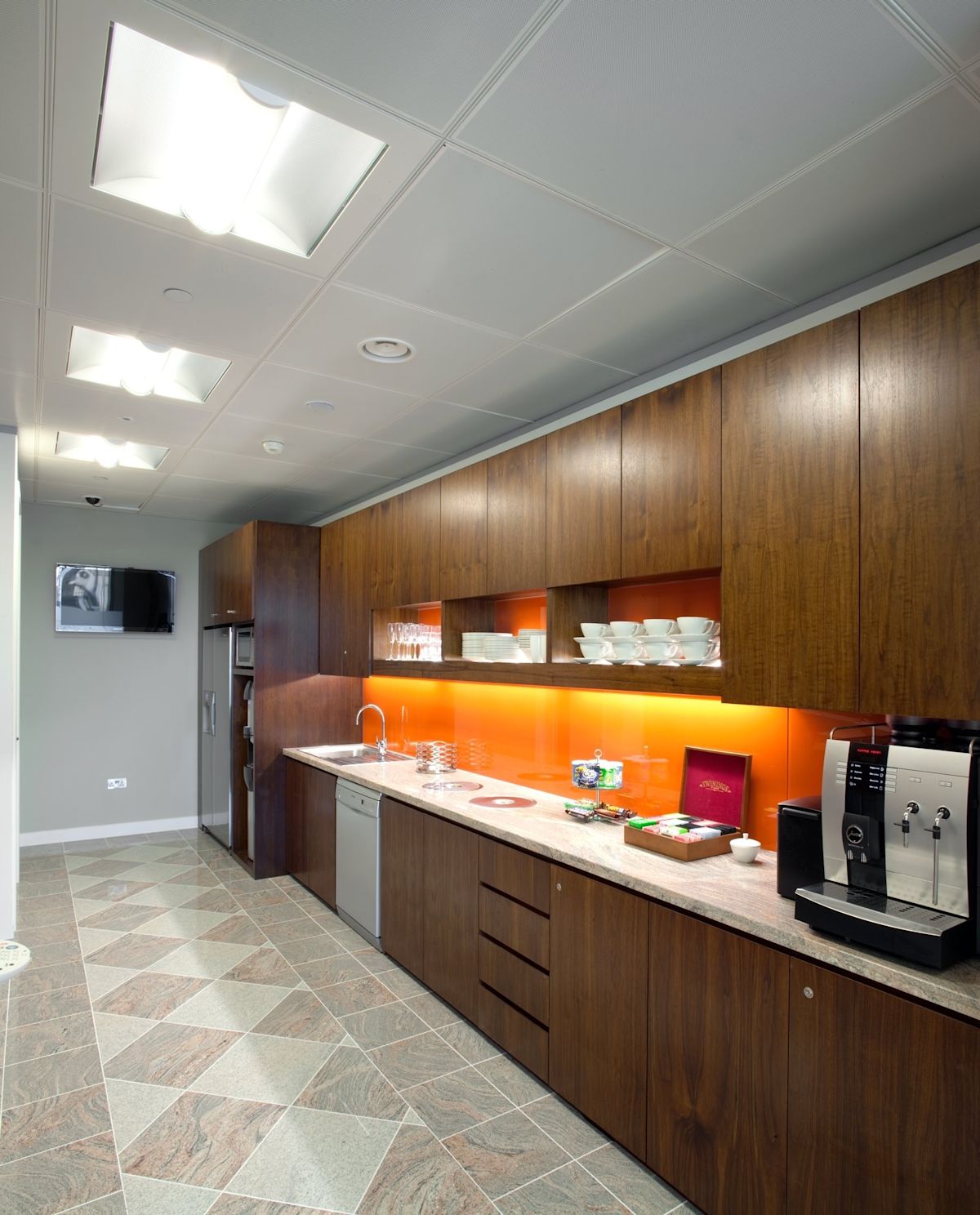 Modern office kitchen with wooden cabinets, a coffee machine, and an orange backsplash.