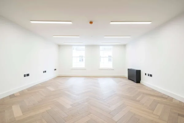 An empty, well-lit room with herringbone parquet flooring and two large windows at Berwick Street 30, London.