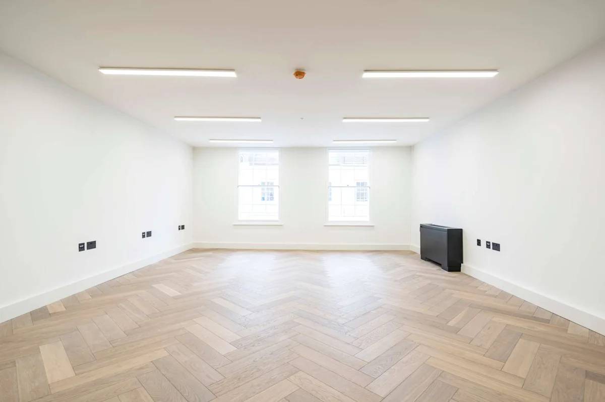 An empty, well-lit room with herringbone parquet flooring and two large windows at Berwick Street 30, London.