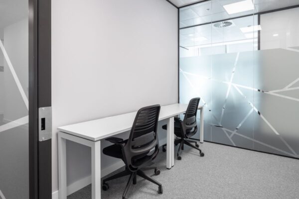 A small, modern office space with two desks and chairs, featuring frosted glass walls with geometric patterns.