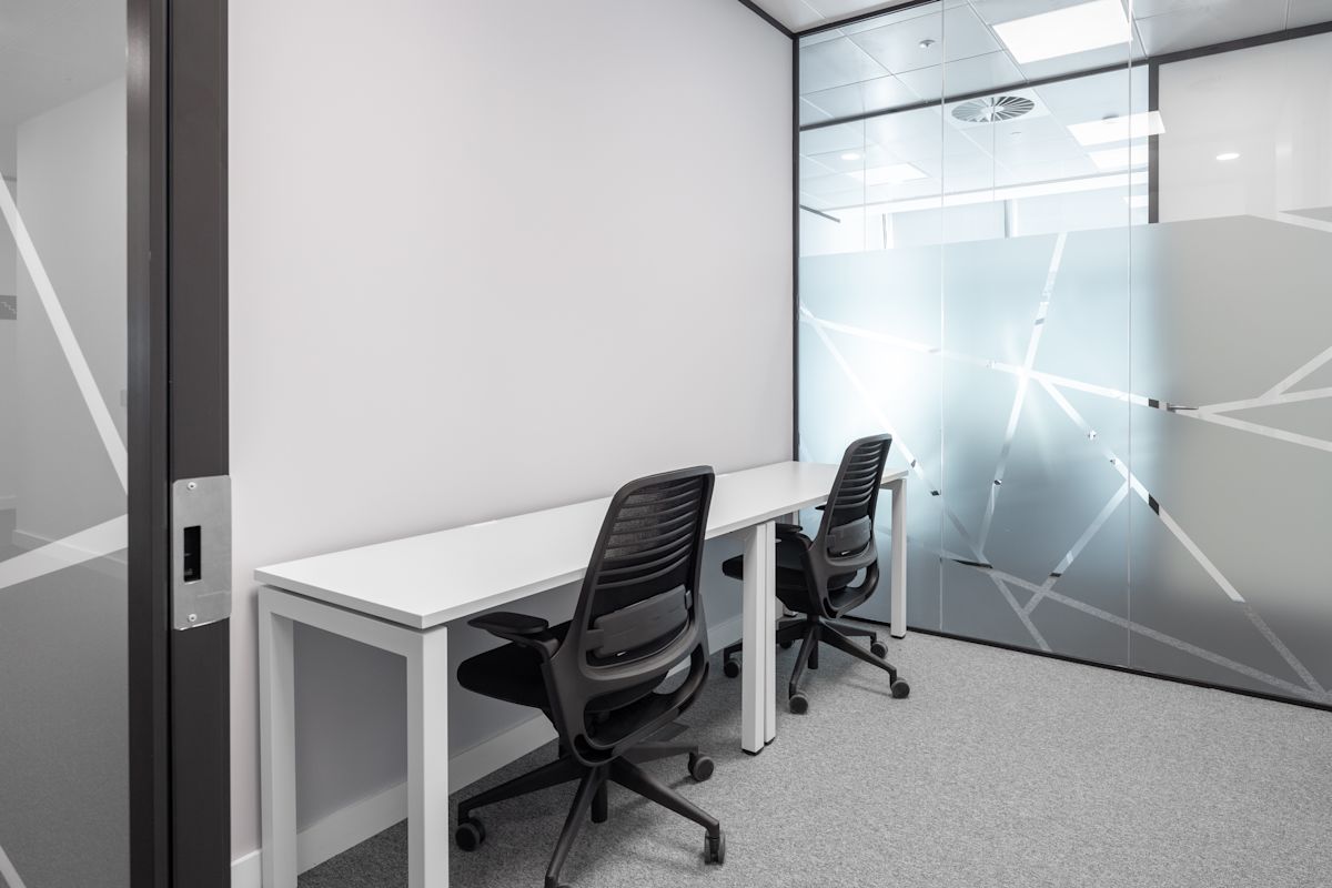 A small, modern office space with two desks and chairs, featuring frosted glass walls with geometric patterns.