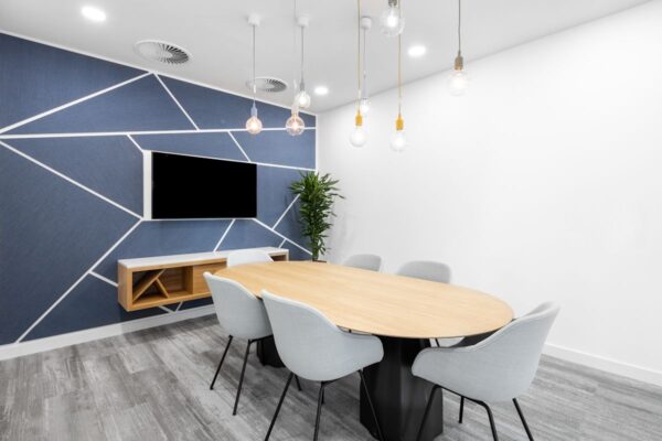 A modern conference room at 100 Bishopsgate, London, featuring a wooden table, grey chairs, geometric wall design, and hanging light bulbs.