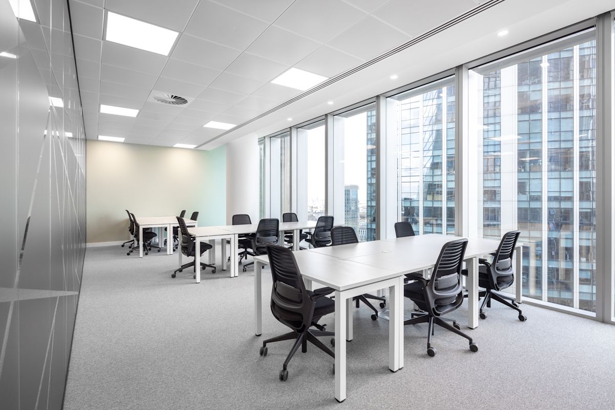A modern office space with several desks and chairs, featuring large windows with a view of adjacent buildings.