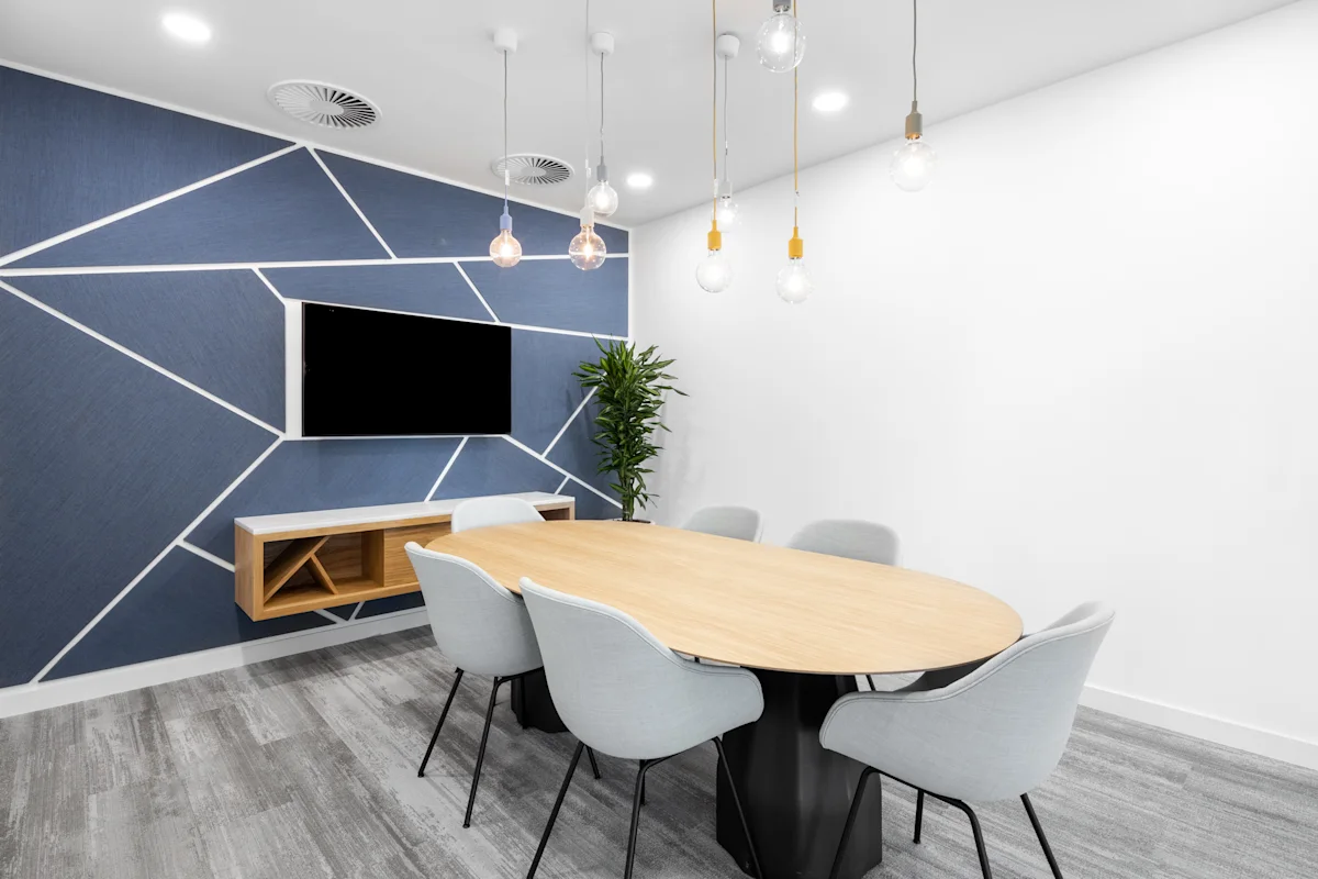 A modern conference room with a wooden oval table, gray chairs, a geometric blue accent wall, and hanging light bulbs.