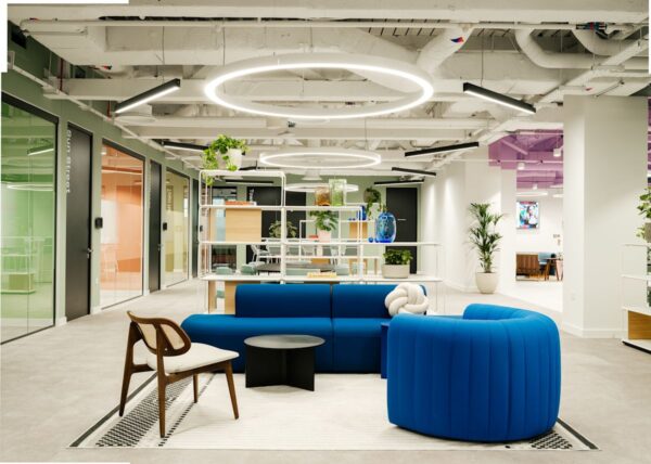 A modern office lounge area with blue sofas, a wooden chair, circular ceiling lights, and plants, at Bishopsgate 199 London.