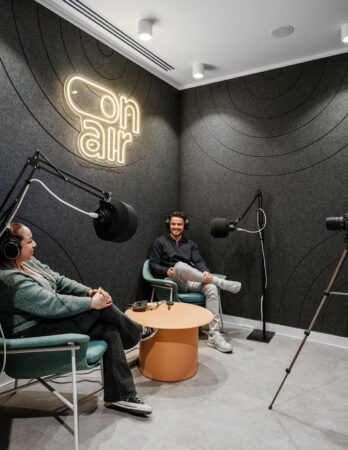 Two people with headphones are sitting in a podcast studio with "on air" sign on the wall.