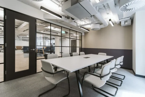 A modern conference room with a long table, surrounded by gray chairs and enclosed by glass walls.
