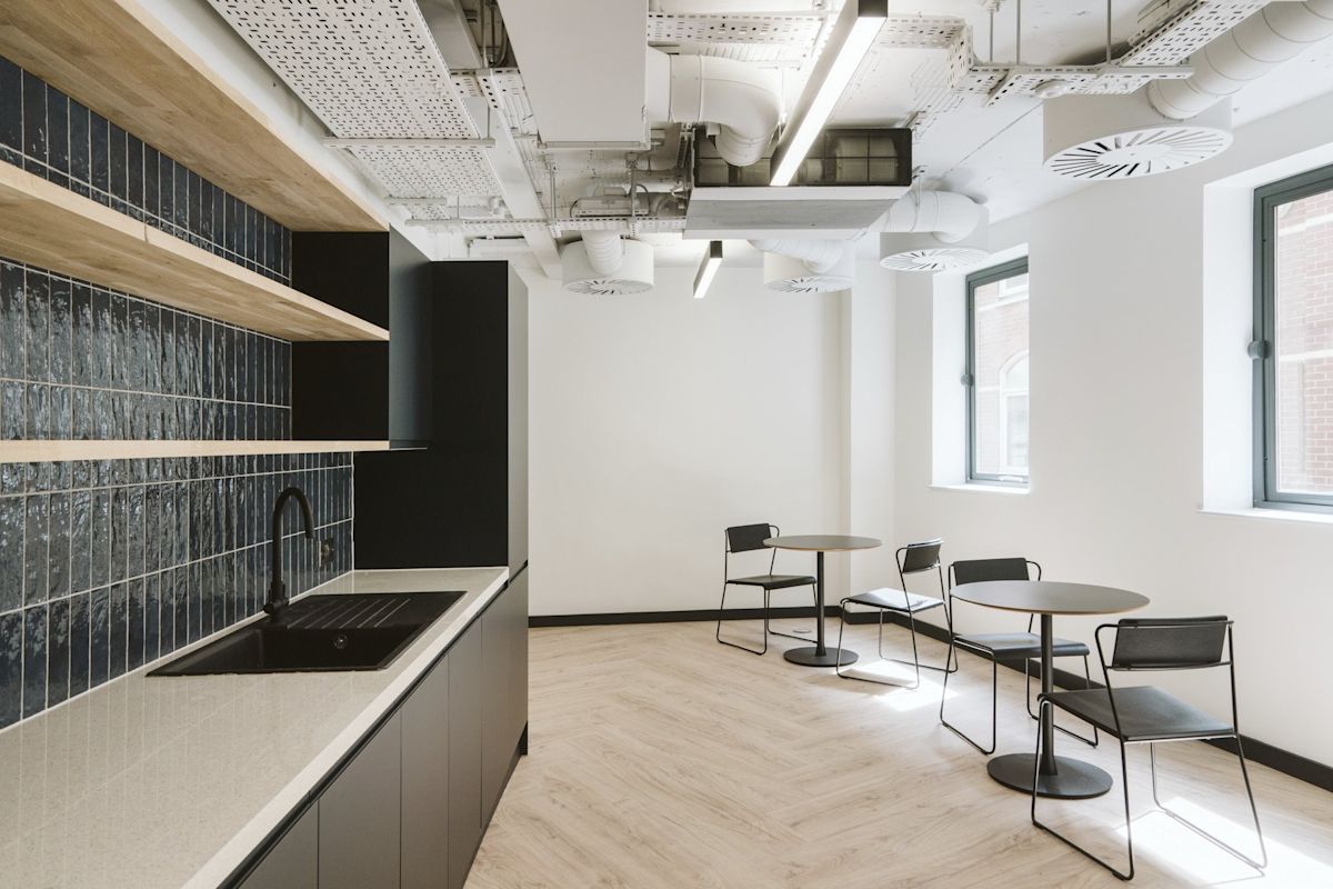A modern office kitchen area with sleek black cabinets, a countertop sink, and small round tables with chairs.