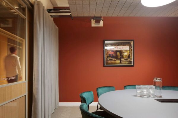 A modern meeting room with a round table, teal chairs, and a red accent wall featuring a framed artwork.