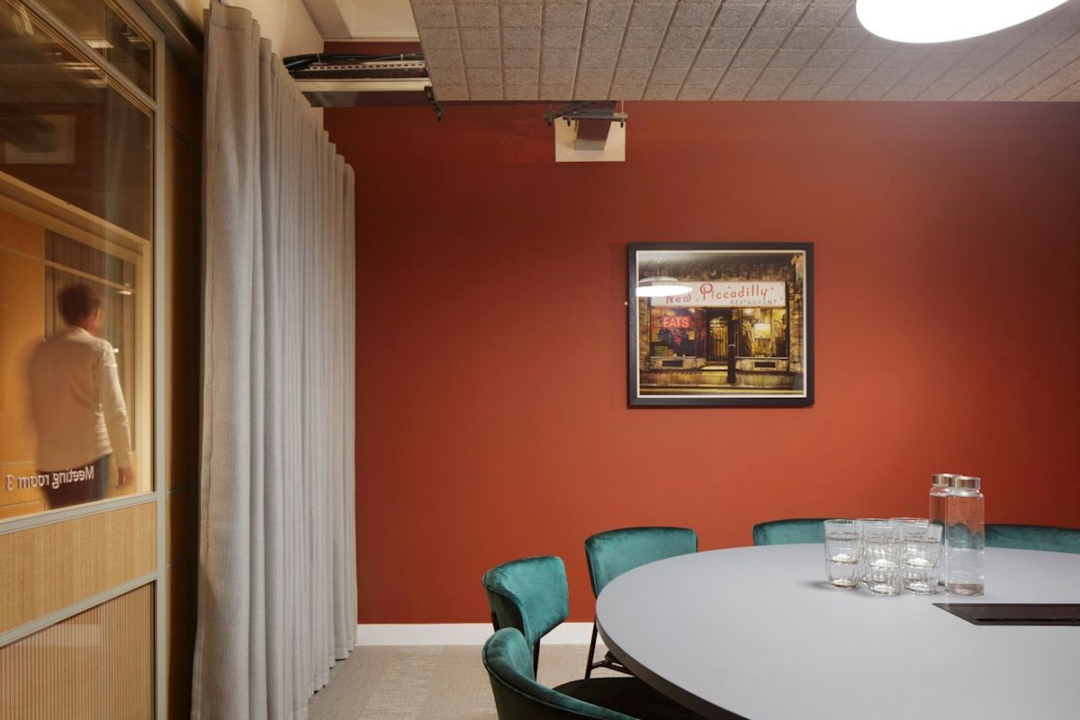 A modern meeting room with a round table, teal chairs, and a red accent wall featuring a framed artwork.
