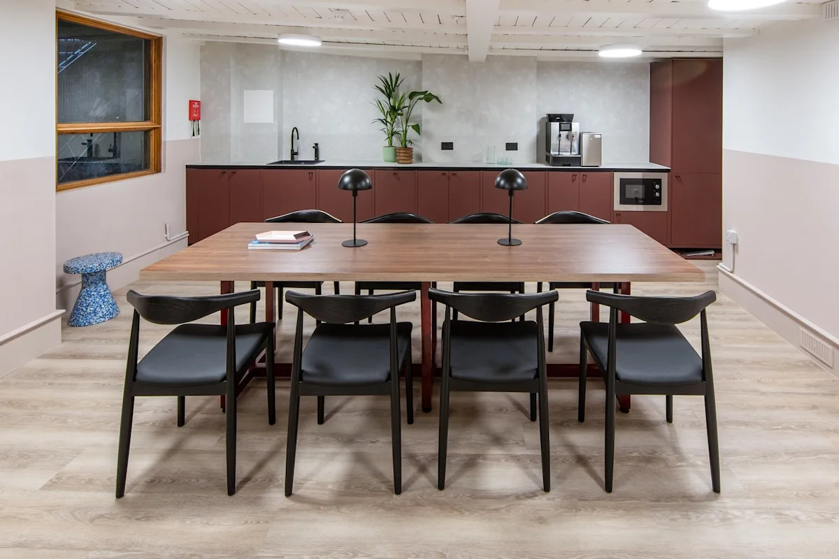 Modern office kitchen with a wooden table, black chairs, and a kitchenette featuring cabinets and appliances.
