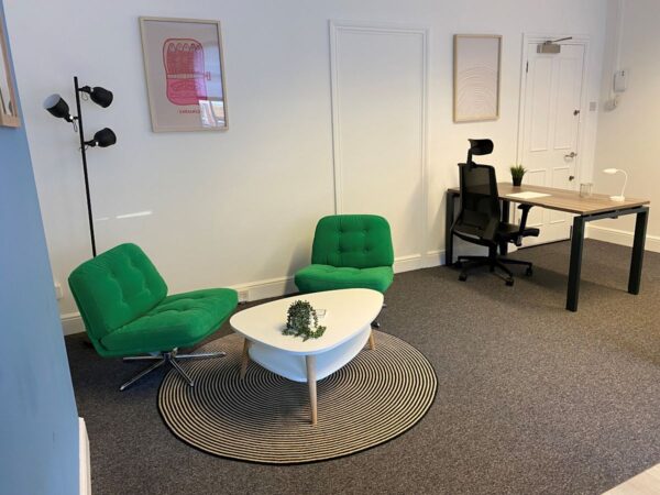 A modern office space with green chairs, a white coffee table on a striped rug, and a desk with a black chair.