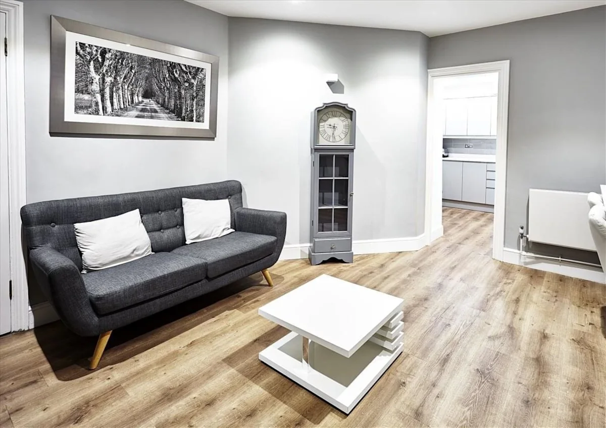 A cozy living room with a gray sofa, white coffee table, wall-mounted photograph, and a tall clock cabinet, featuring light wooden flooring.