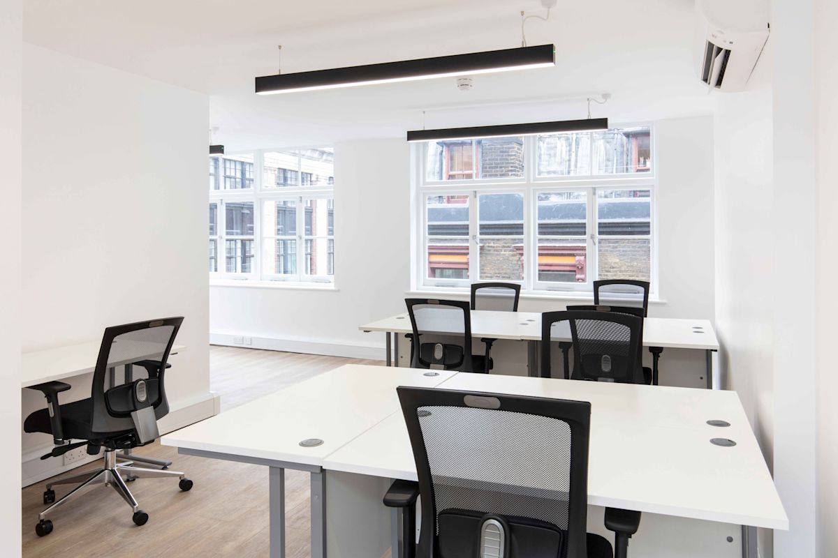 A modern office space with several white desks and black chairs, featuring large windows and wooden floors.