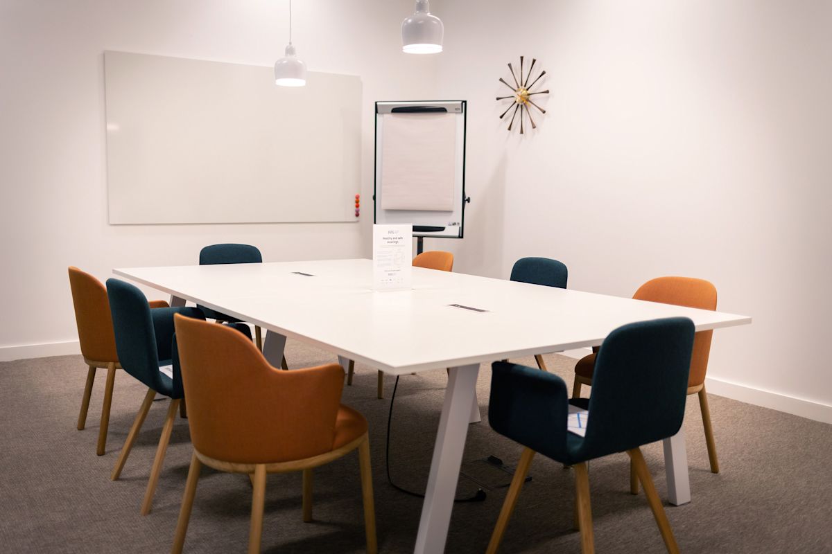 A modern meeting room with a large white table surrounded by colorful chairs, a whiteboard, and a flip chart.