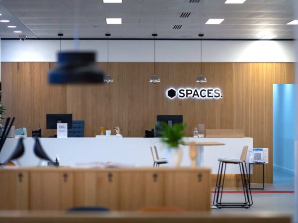 A modern office reception area with a wooden backdrop and the "Spaces" logo illuminated on the wall.