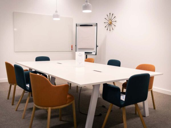 A modern conference room at Cabot Square 25, London, featuring a white table with multicolored chairs and a flip chart.