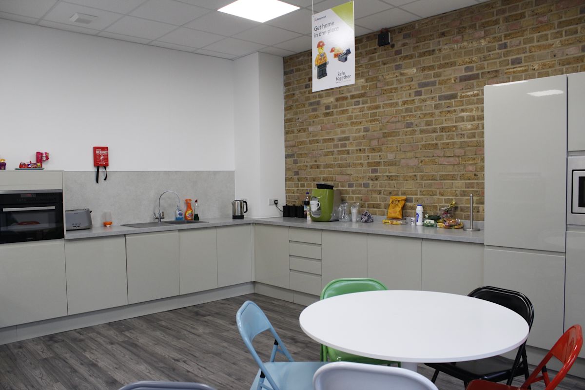 Modern office kitchen with a brick accent wall, table, and colorful chairs.