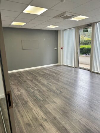 An empty office space on the ground floor of Cadmus Court in London, featuring wooden flooring and large windows with vertical blinds.