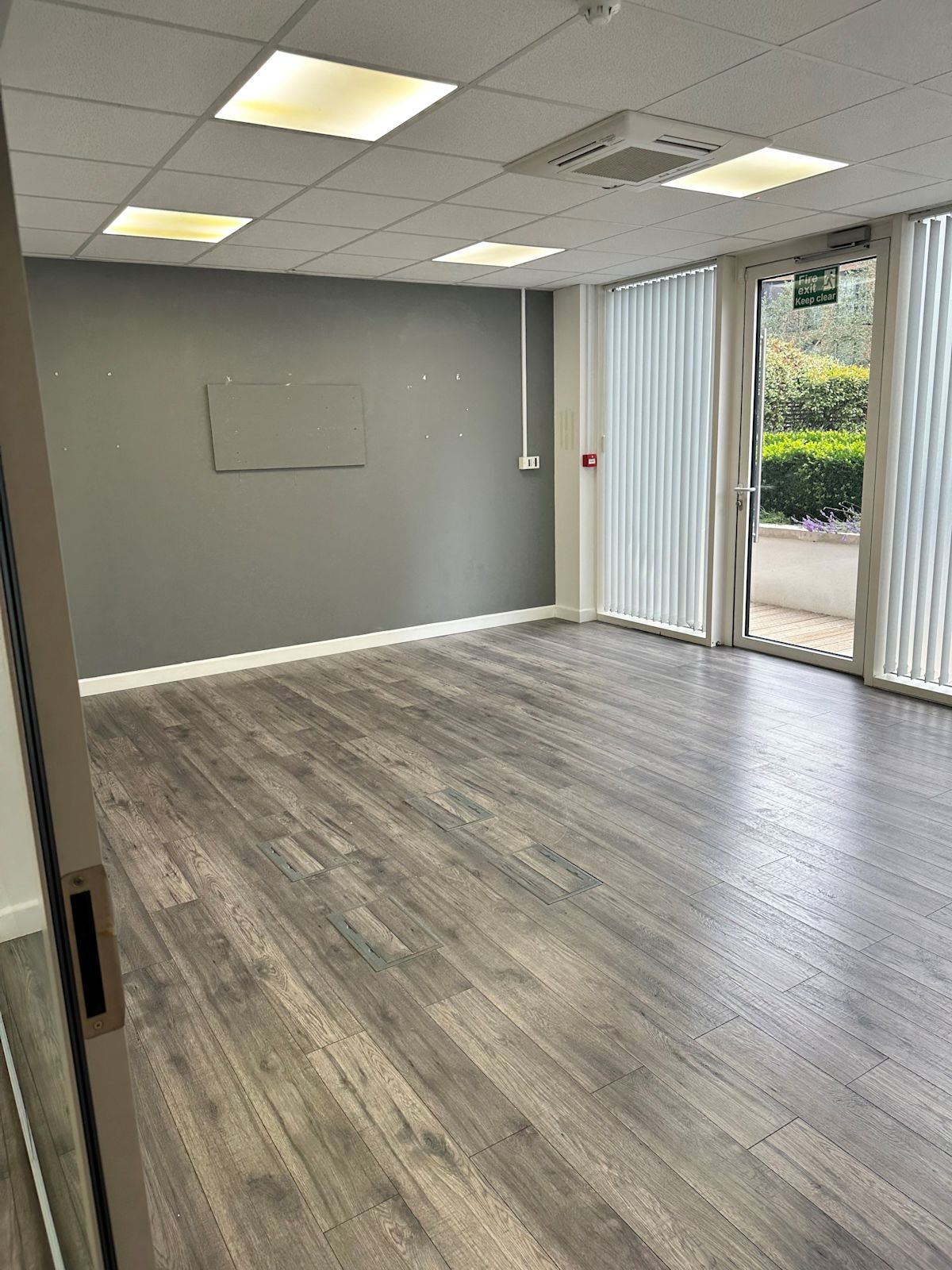 An empty office space on the ground floor of Cadmus Court in London, featuring wooden flooring and large windows with vertical blinds.
