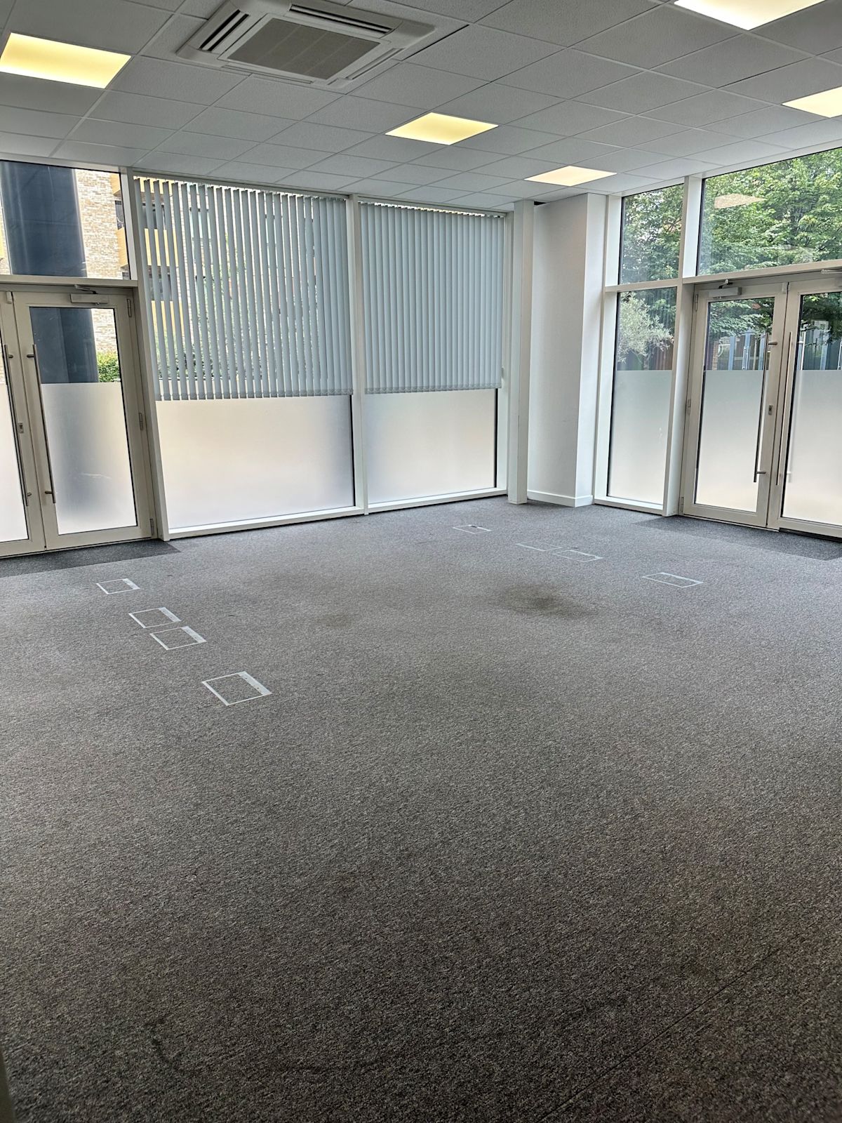 Spacious, empty office space with large windows and gray carpeting on the ground floor of Cadmus Court in London.