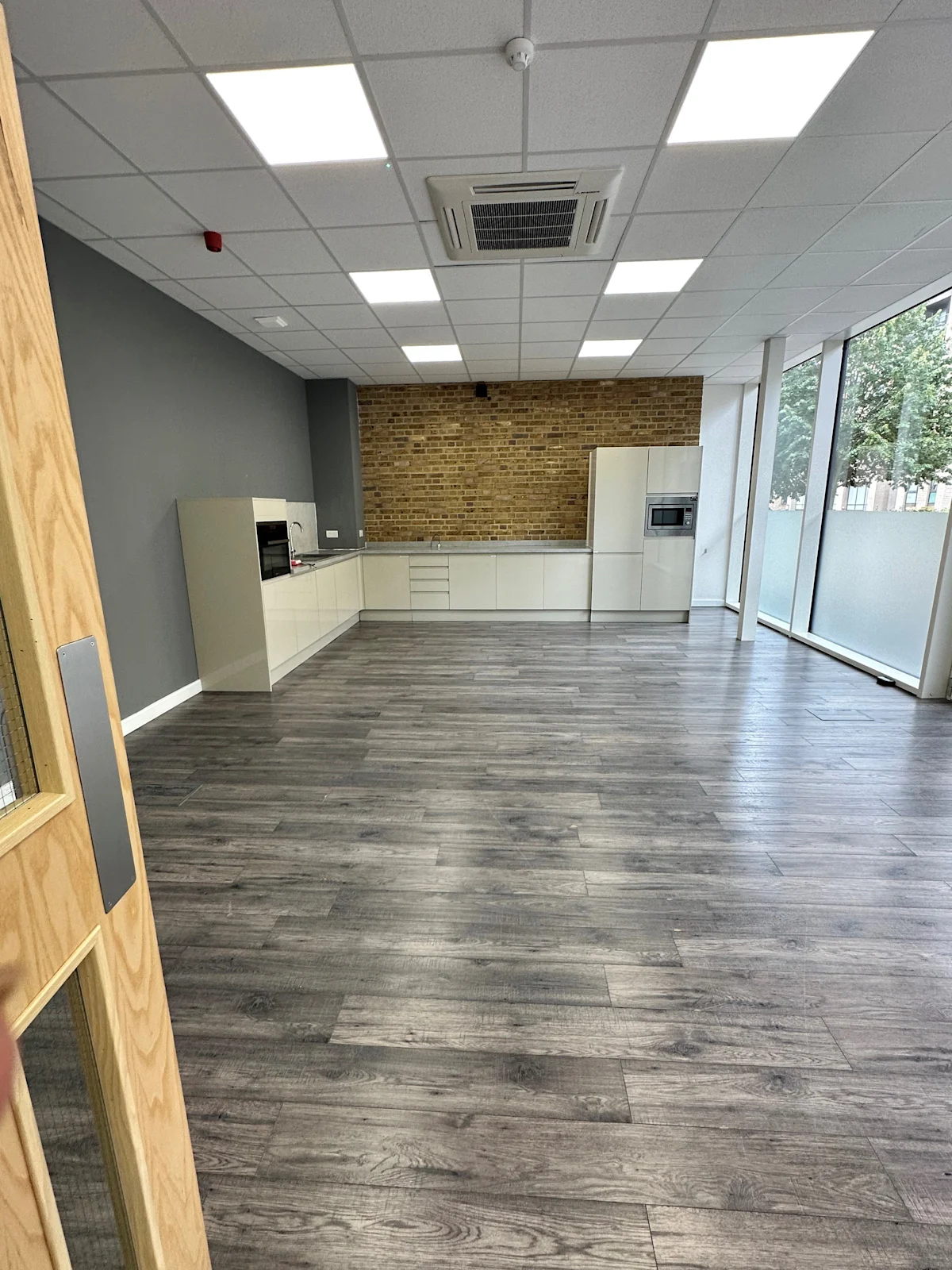 A modern, empty office space with sleek wooden floors and a kitchenette area, located on the ground floor of Cadmus Court, London.