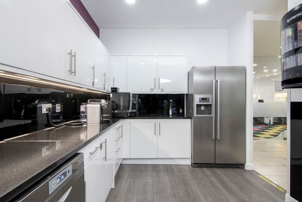 A modern office kitchen with white cabinets, a stainless steel fridge, and coffee machines.