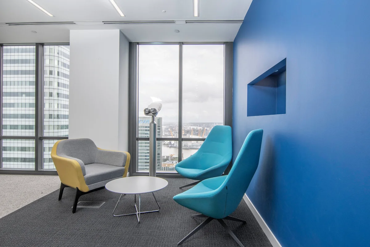 Modern office seating area in Canada Square 1, London, with blue and yellow chairs, a small table, and a large window view.