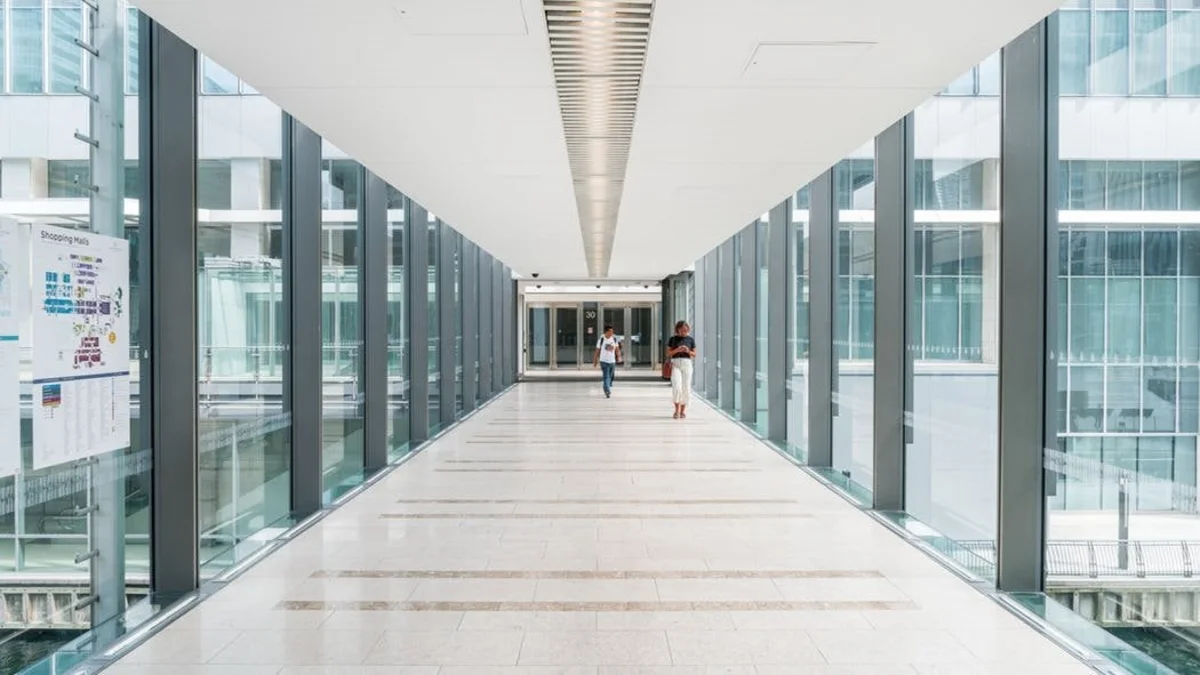 A modern glass walkway inside Churchill Place 30 in London, with two people walking towards the exit.